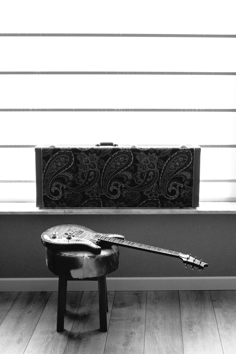 An electric guitar resting on a stool below a window, its case on the sill behind